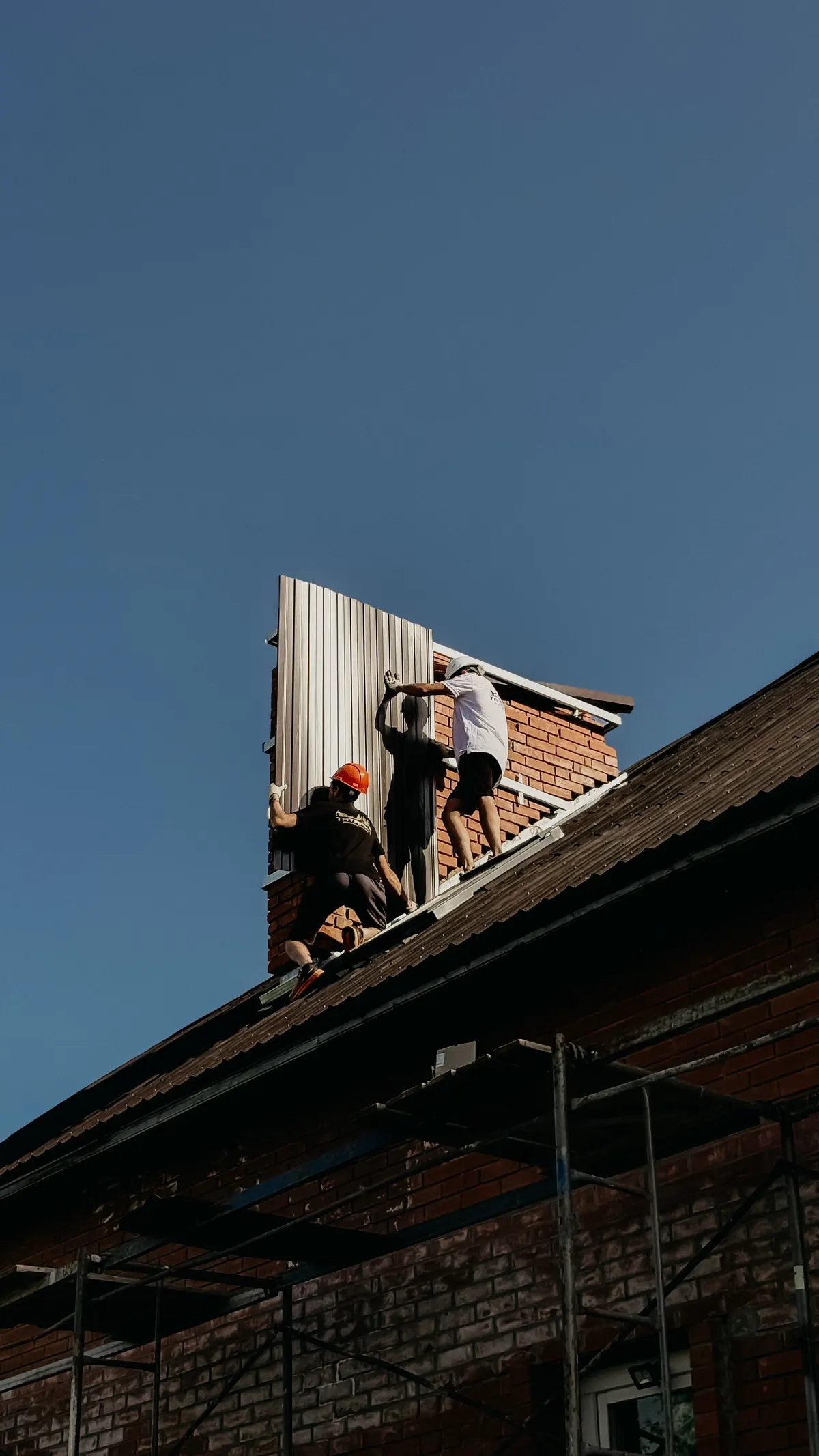 Workers installing metal roof panels on a brick house